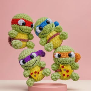 以下が含まれることがあります： 手作りの編みぐるみ忍者タートルズ4体。各タートルズは異なる色のマスクをしています：赤、青、紫、オレンジ。彼らは緑色で黄色いお腹をしており、ピザのスライスを持っています。タートルズはピンク色のプラットフォームに展示されています。