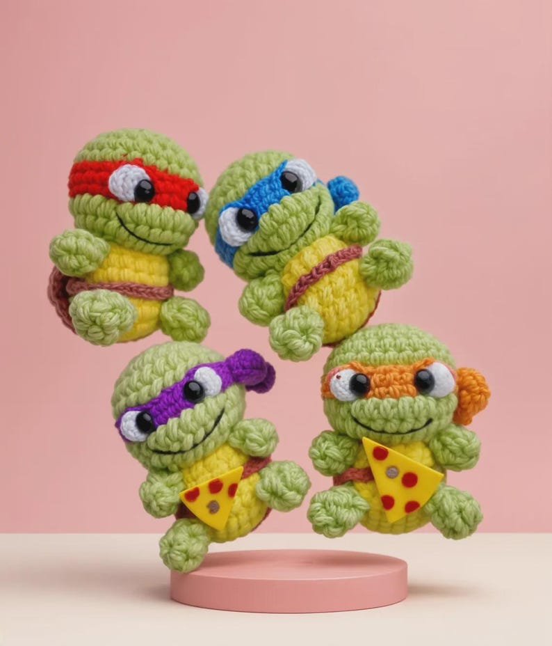 May include: Four handmade crochet Teenage Mutant Ninja Turtles. Each turtle has a different colored mask: red, blue, purple, and orange. They are green with yellow bellies and hold a slice of pizza. The turtles are displayed on a pink platform.