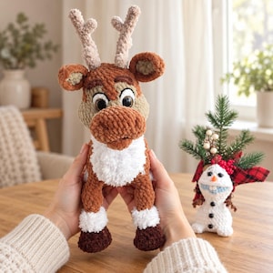 Puede incluir: Un juguete de reno tejido a ganchillo con hilo marrón, beige y blanco, con grandes ojos oscuros y astas texturizadas. A su lado hay una pequeña figura de muñeco de nieve tejida en blanco con nariz de zanahoria y botones negros decorativos.