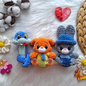 Op de afbeelding: Drie handgemaakte gehaakte knuffels: een blauwe slang, een oranje hond en een grijze konijn met een blauwe hoed. Het speelgoed ligt op een witte, zachte ondergrond, met decoratieve bloemen en een hartvormig ornament.