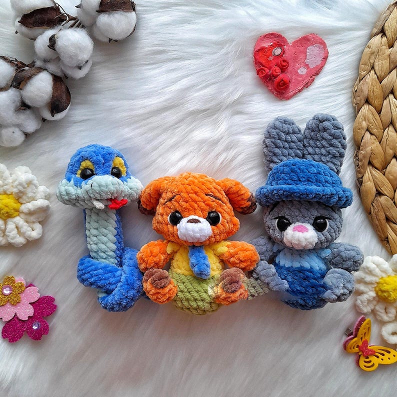 Op de afbeelding: Drie handgemaakte gehaakte knuffels: een blauwe slang, een oranje hond en een grijze konijn met een blauwe hoed. Het speelgoed ligt op een witte, zachte ondergrond, met decoratieve bloemen en een hartvormig ornament.