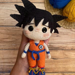 May include: A handmade crochet doll of a character with spiky black hair, wearing an orange jumpsuit with a blue belt and blue shoes with yellow accents. The doll has large black eyes and a small patch on the chest.
