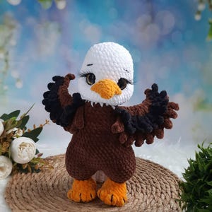 May include: A crocheted bald eagle plush toy featuring a white head, a bright yellow beak, and a brown body with dark brown wings extended outward. The eagle has large, dark, glossy eyes with long black eyelashes and textured orange feet.