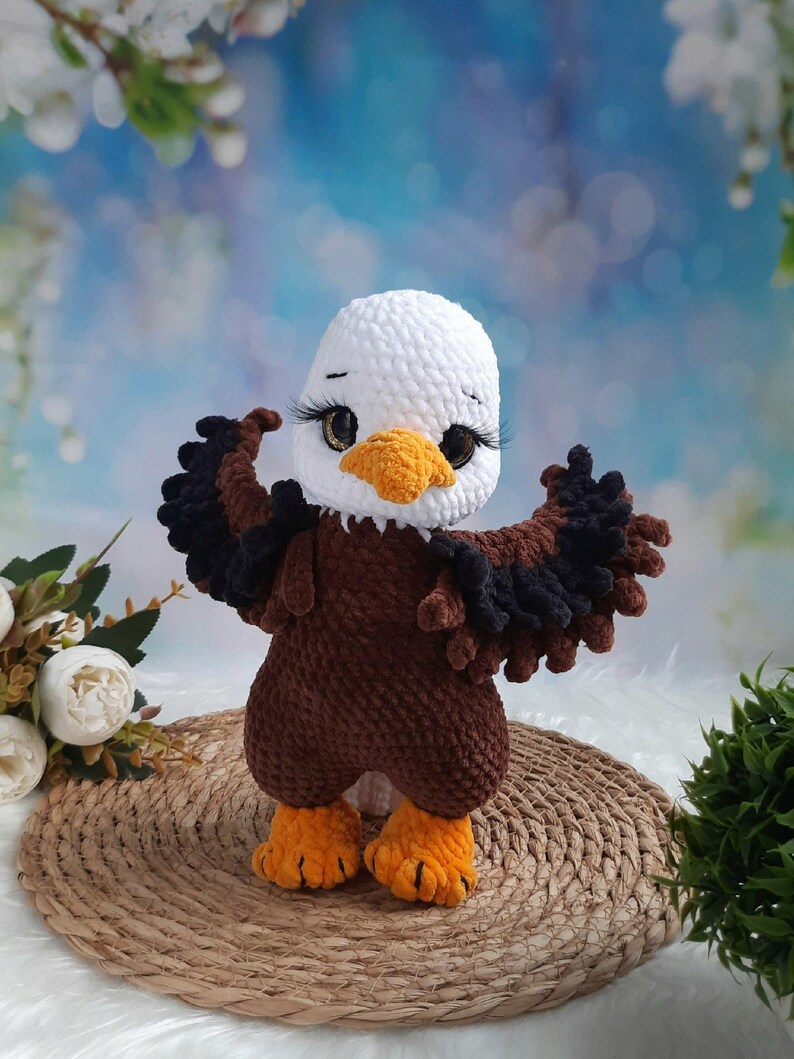 May include: A crocheted bald eagle plush toy featuring a white head, a bright yellow beak, and a brown body with dark brown wings extended outward. The eagle has large, dark, glossy eyes with long black eyelashes and textured orange feet.