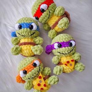 May include: Four handmade crochet Teenage Mutant Ninja Turtles. Each turtle has a different colored mask: red, blue, purple, and orange. They have yellow shells and brown belts. Two turtles hold a felt pizza slice.