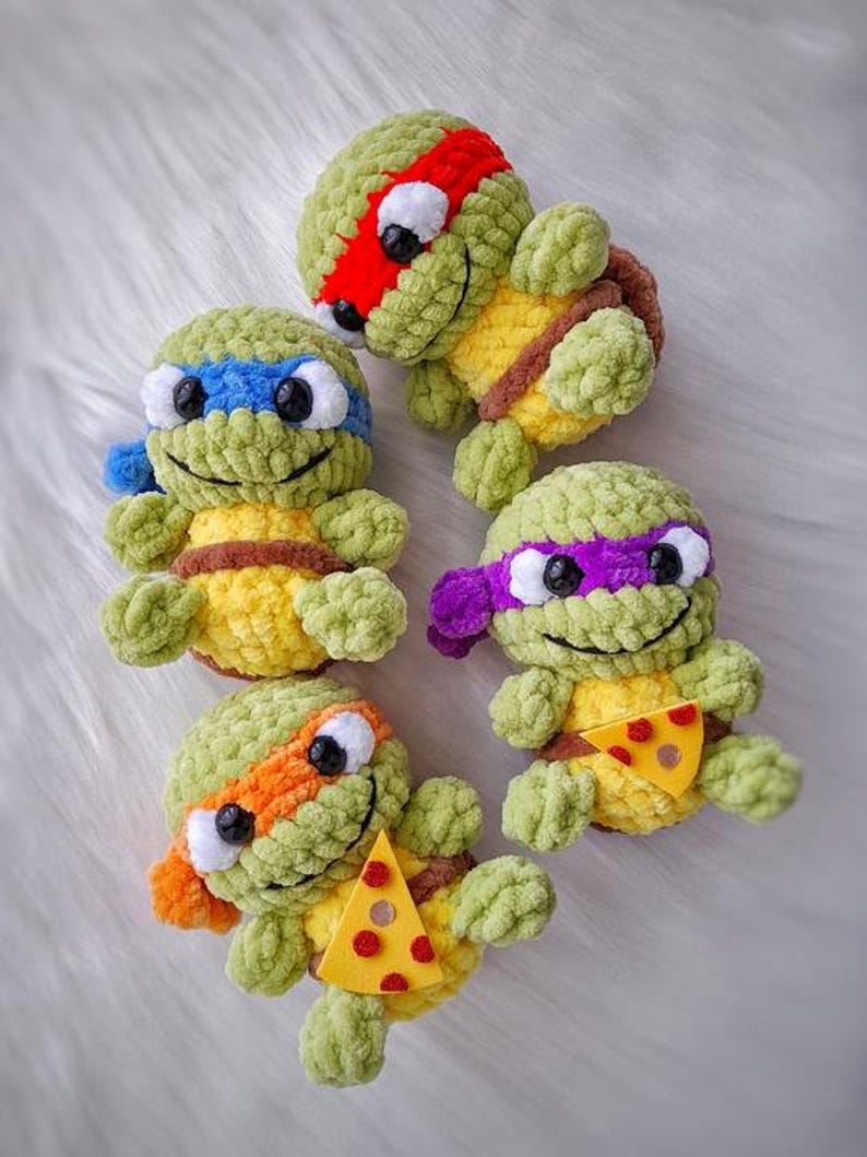 May include: Four handmade crochet Teenage Mutant Ninja Turtles. Each turtle has a different colored mask: red, blue, purple, and orange. They have yellow shells and brown belts. Two turtles hold a felt pizza slice.