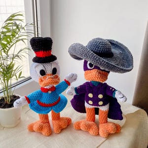 May include: Two handmade crochet duck dolls. One duck wears a black top hat with a red band, a blue outfit, and a red bow tie. The other duck is dressed in a purple coat, a gray hat, and a purple mask. Both have orange feet.
