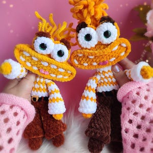 Puede incluir: Dos muñecos de ganchillo hechos a mano con ojos grandes y expresivos y pelo amarillo. Los muñecos tienen brazos a rayas amarillas y blancas, pantalones marrones y detalles amarillos y blancos. Los muñecos se sostienen sobre un fondo rosa.