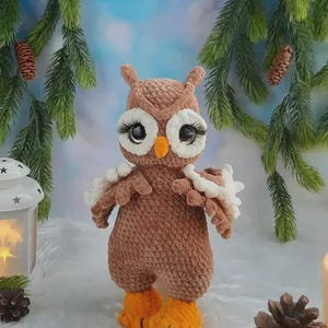 May include: A brown crocheted owl plush toy with large black eyes, long black eyelashes, white eye patches, and a small orange beak. The owl has textured brown yarn wings with white accents and bright orange feet, standing upright.