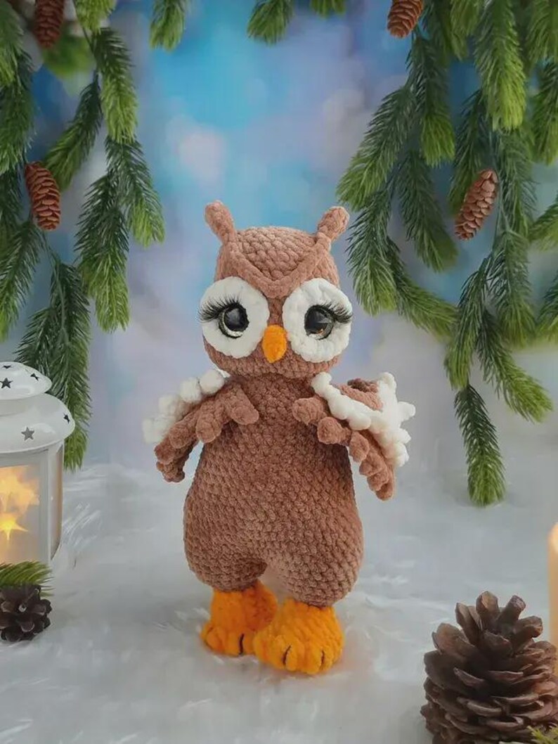 May include: A brown crocheted owl plush toy with large black eyes, long black eyelashes, white eye patches, and a small orange beak. The owl has textured brown yarn wings with white accents and bright orange feet, standing upright.
