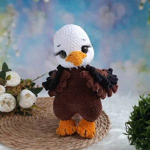 May include: A crocheted bald eagle plush toy featuring a white head, a brown body, and bright orange feet and beak. The eagle has large, dark eyes with long, prominent eyelashes, and textured, dark brown and black wing details on its sides.