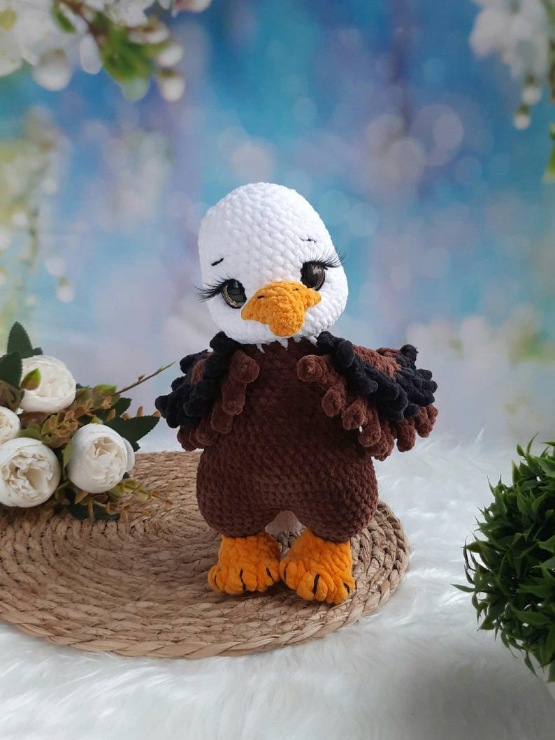 May include: A crocheted bald eagle plush toy featuring a white head, a brown body, and bright orange feet and beak. The eagle has large, dark eyes with long, prominent eyelashes, and textured, dark brown and black wing details on its sides.
