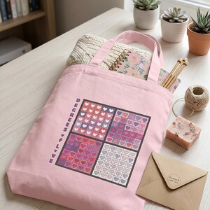 May include: A light pink canvas tote bag featuring a square graphic divided into four quadrants filled with small hearts in shades of pink, purple, and white. The text DEGREES OF LOVE is printed vertically in purple letters along the left side.