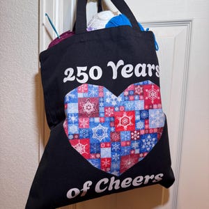 May include: A black tote bag with two handles, featuring the white text 250 Years of Cheers above and below a large heart graphic. The heart is composed of a patchwork of red, white, and blue squares, each decorated with various white snowflake patterns.