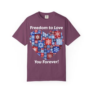 Peut inclure: Un t-shirt à manches courtes de couleur prune avec un grand motif en forme de cœur composé d'un patchwork de flocons de neige rouges, blancs et bleus. Le texte "Freedom to Love" est inscrit au-dessus et "You Forever!" en dessous en blanc.