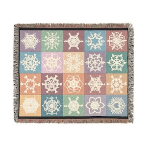 Peut inclure: Une couverture tissée avec un motif de grille de carrés, chacun présentant un design de flocon de neige blanc unique. La couverture a un bord à franges et une variété de couleurs pastel, dont le violet, le bleu, le vert et l'orange.
