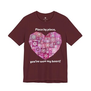 以下が含まれることがあります： マルーンカラーの半袖Tシャツ。ピンクと白の様々な雪の結晶模様で構成された大きなハートのグラフィックが特徴です。ハートのデザインの上下には「Piece by piece, you've won my heart!」という白い文字がプリントされています。