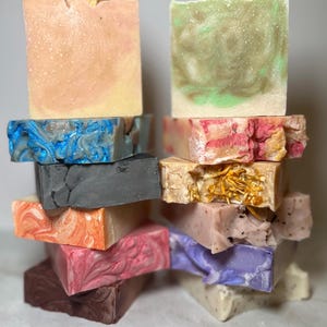 Op de afbeelding: Een stapel handgemaakte zepen in verschillende kleuren en ontwerpen. De zepen bevatten tinten roze, oranje, blauw, groen en bruin. Sommige zepen hebben decoratieve elementen zoals gedroogde sinaasappelschijfjes of bloemaccenten. De zepen zijn op elkaar gestapeld.