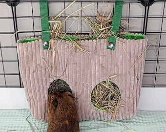 May include: A brown and white guinea pig is sticking its head into a hanging hay feeder. The feeder is made of brown corduroy with green trim and has two openings. The feeder is hanging from a black wire cage.