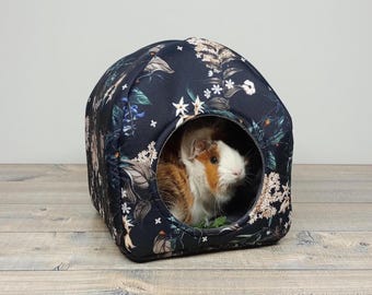 May include: A small pet house with a floral pattern in navy blue, white, and green. A guinea pig with brown, white, and orange fur is inside the house, looking out of the round opening. The house is on a wooden surface.