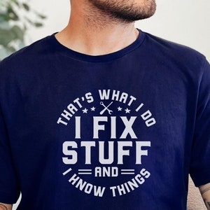 May include: Navy blue t-shirt with the text "THAT'S WHAT I DO, I FIX STUFF AND I KNOW THINGS" in white. The shirt has a crew neck and short sleeves. A red tag indicates sizes 3XL-4XL-5XL are available.