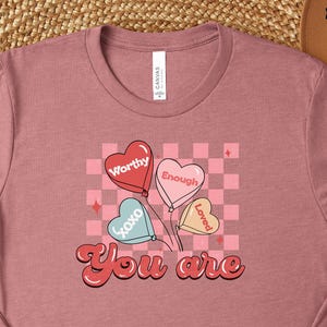 May include: Heather mauve t-shirt with a graphic of heart-shaped balloons that read "Worthy," "Enough," and "Loved." The text "You are" is below the balloons. The shirt is available in sizes 3XL-5XL.