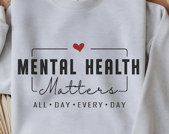 May include: A light gray sweatshirt with the text "MENTAL HEALTH Matters" in black, with a red heart above the word "Matters". The phrase "ALL DAY • EVERY DAY" is below. A tan hat and woven placemats are in the background.