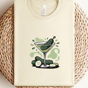 May include: A natural-colored t-shirt featuring a cartoon illustration of a martini glass with a pickle garnish. Additional pickles and green accents complete the design. The word "NATURAL" is printed at the bottom.