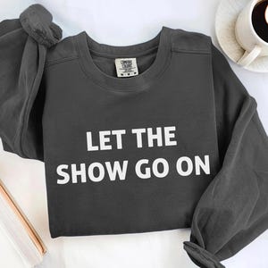 May include: Dark gray sweatshirt with the text "LET THE SHOW GO ON" in white block letters. The sweatshirt has long sleeves and a crew neck. A coffee cup and a book are in the background.