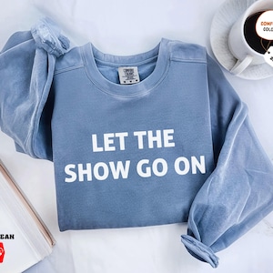 May include: A blue jean-colored sweatshirt with the text "LET THE SHOW GO ON" in white block letters. The sweatshirt has long sleeves and a crew neck. A coffee cup and a notepad are in the background.
