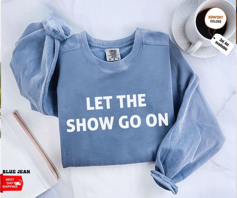 May include: A blue jean-colored sweatshirt with the text "LET THE SHOW GO ON" in white block letters. The sweatshirt has long sleeves and a crew neck. A coffee cup and a notepad are in the background.