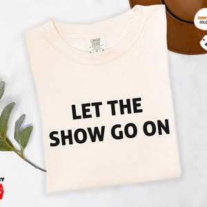 May include: Ivory-colored t-shirt with the text "LET THE SHOW GO ON" in black. The shirt is a Comfort Colors brand. A brown hat and a sprig of greenery are in the background.