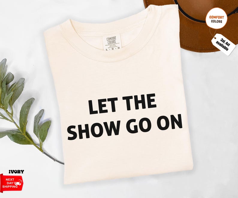 May include: Ivory-colored t-shirt with the text "LET THE SHOW GO ON" in black. The shirt is a Comfort Colors brand. A brown hat and a sprig of greenery are in the background.