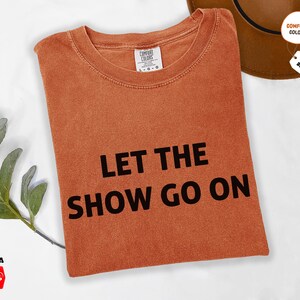 May include: An orange Comfort Colors t-shirt with the phrase "LET THE SHOW GO ON" in bold black letters. The shirt is folded and has a tag indicating available sizes. A brown hat is partially visible.