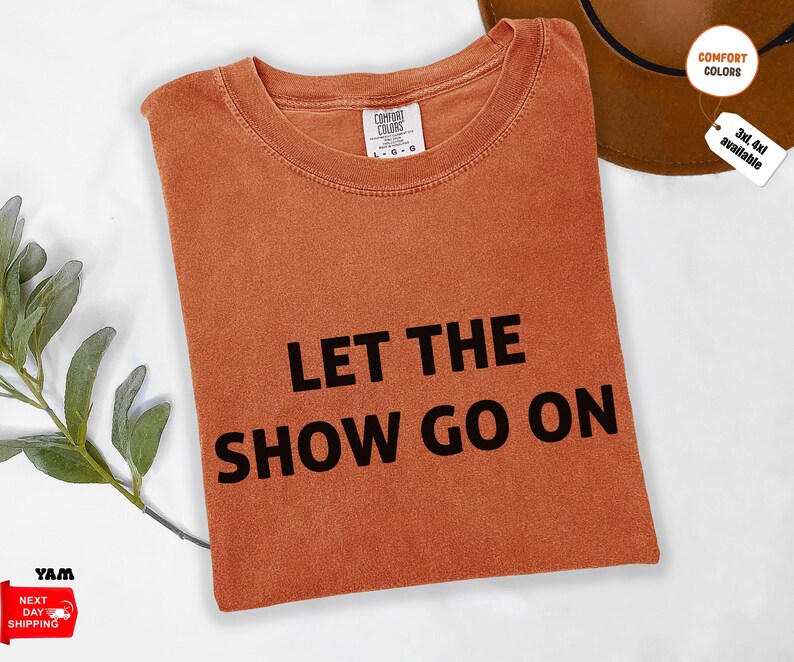 May include: An orange Comfort Colors t-shirt with the phrase "LET THE SHOW GO ON" in bold black letters. The shirt is folded and has a tag indicating available sizes. A brown hat is partially visible.