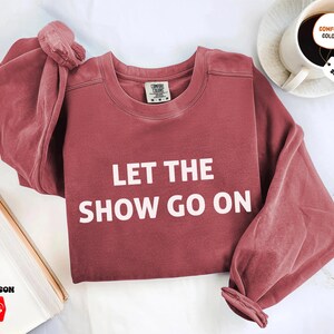 May include: A maroon-colored sweatshirt with the text "LET THE SHOW GO ON" in white block letters. The sweatshirt has long sleeves and a crew neck. The image also shows a coffee cup and a book.