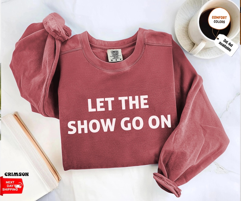 May include: A maroon-colored sweatshirt with the text "LET THE SHOW GO ON" in white block letters. The sweatshirt has long sleeves and a crew neck. The image also shows a coffee cup and a book.
