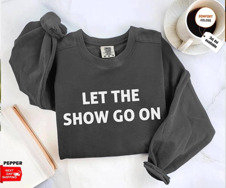 May include: Dark gray sweatshirt with the text "LET THE SHOW GO ON" in white block letters. The sweatshirt has long sleeves and a crew neck. A coffee cup and a book are in the background.