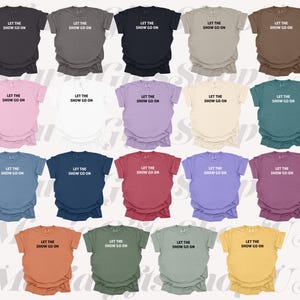 May include: A collection of short-sleeved t-shirts in various colors, each displaying the text "LET THE SHOW GO ON" in a simple, white font. The shirts are arranged in rows, showcasing a range of hues from dark to light.