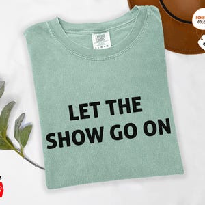 May include: A light green t-shirt with the text "LET THE SHOW GO ON" in black. The shirt is folded and has a tag that says "COMFORT COLORS". A brown hat is in the background.