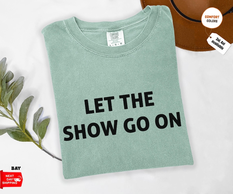 May include: A light green t-shirt with the text "LET THE SHOW GO ON" in black. The shirt is folded and has a tag that says "COMFORT COLORS". A brown hat is in the background.