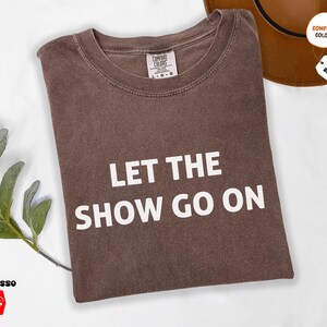 May include: Espresso-colored t-shirt with the text "LET THE SHOW GO ON" in white block letters. The shirt is folded and has a crew neck. A brown hat is visible in the background. The shirt is available in sizes 3XL and 4XL.