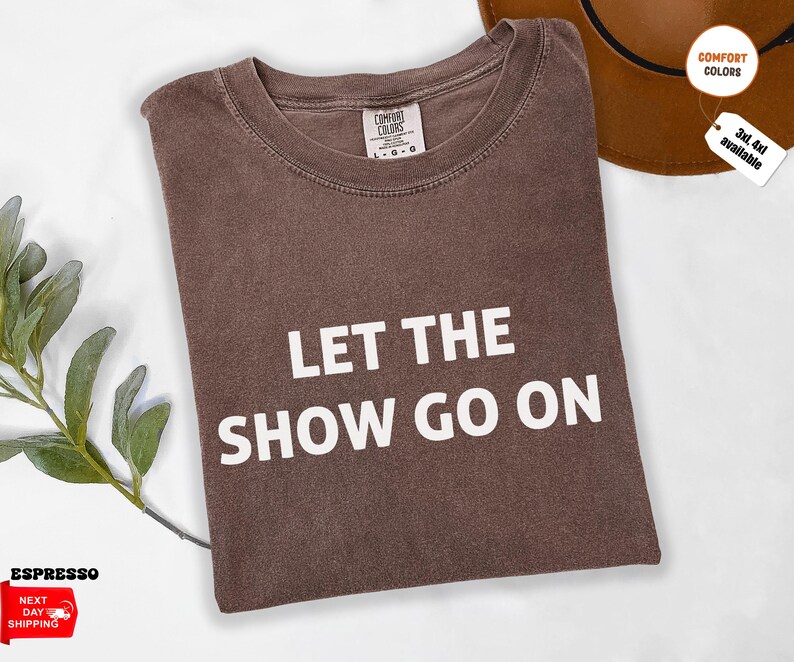 May include: Espresso-colored t-shirt with the text "LET THE SHOW GO ON" in white block letters. The shirt is folded and has a crew neck. A brown hat is visible in the background. The shirt is available in sizes 3XL and 4XL.