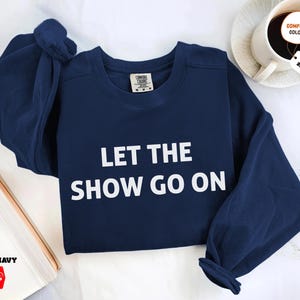 May include: A navy blue sweatshirt with the text "LET THE SHOW GO ON" in white block letters. The sweatshirt has a crew neck and long sleeves. The Comfort Colors brand tag is visible. The image also shows a coffee cup and a book.
