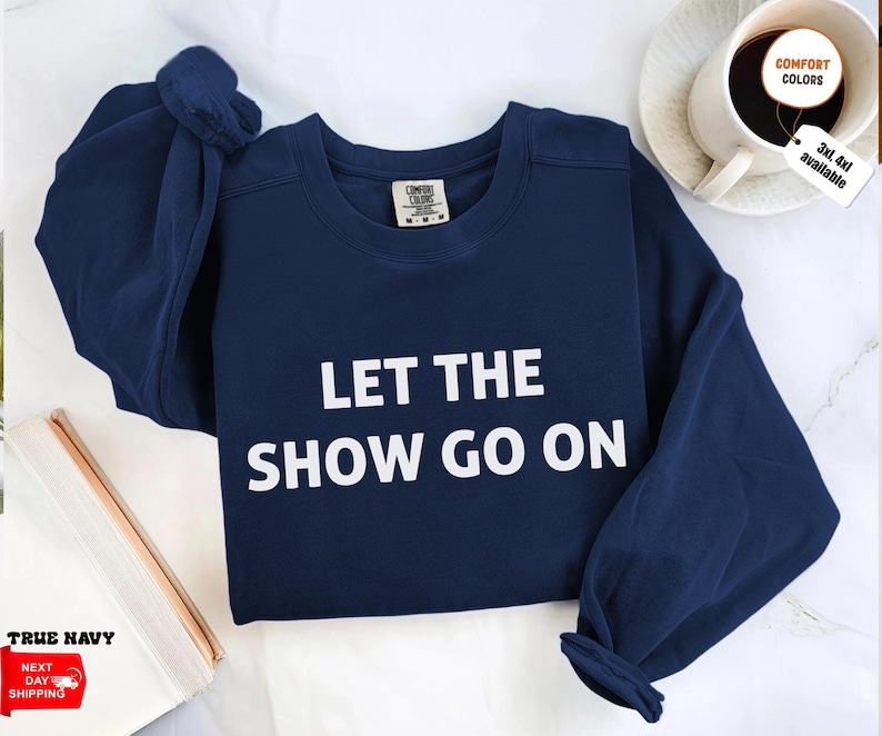 May include: A navy blue sweatshirt with the text "LET THE SHOW GO ON" in white block letters. The sweatshirt has a crew neck and long sleeves. The Comfort Colors brand tag is visible. The image also shows a coffee cup and a book.