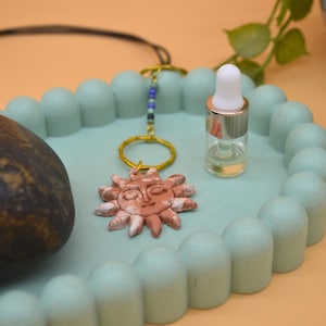 May include: A terracotta-coloured sun pendant with a face, attached to a gold-toned ring and a short strand of blue and green beads on a black cord. Next to it sits a small, clear glass dropper bottle with a white cap and a dark smooth stone.