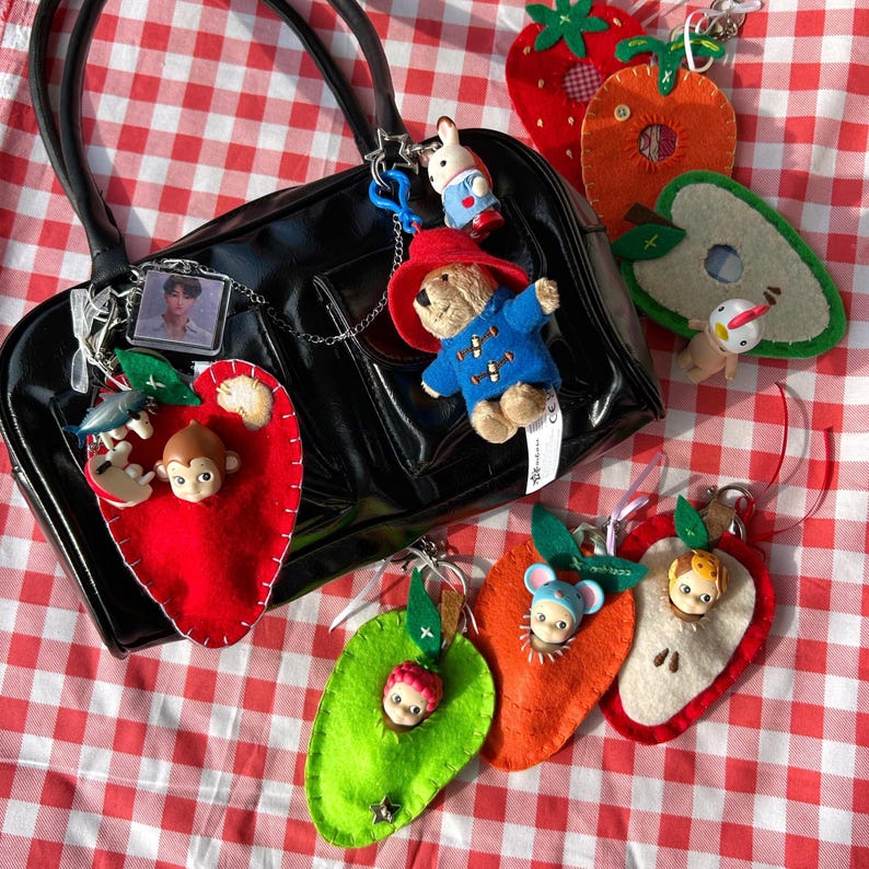 May include: A black handbag with multiple keyrings attached. Keyrings feature a Paddington Bear, a bunny, and felt fruit shapes with small figurines. The background is a red and white gingham pattern.
