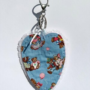 May include: Heart-shaped keyring with a blue fabric patterned with teddy bears in outfits. The heart is edged with white lace and attached to a silver-coloured clasp and ring. A charming and whimsical accessory.