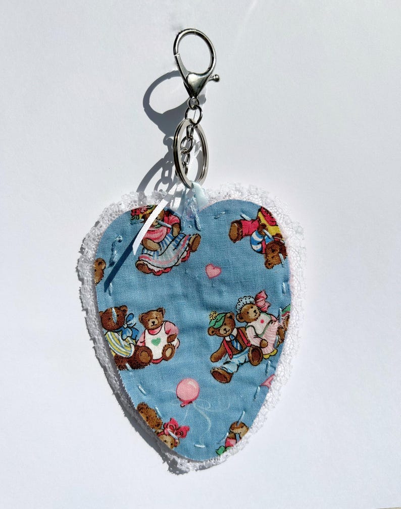 May include: Heart-shaped keyring with a blue fabric patterned with teddy bears in outfits. The heart is edged with white lace and attached to a silver-coloured clasp and ring. A charming and whimsical accessory.