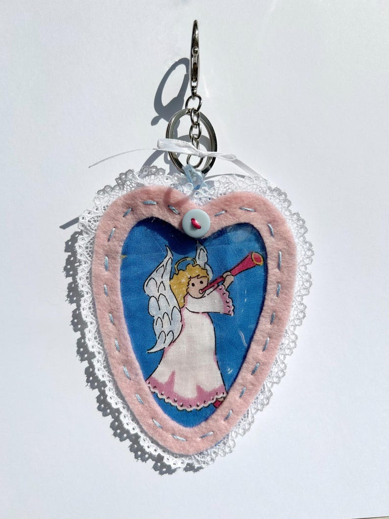 May include: A heart-shaped keyring with a pink felt border and a blue fabric centre. The centre features a cartoon angel playing a trumpet. The keyring has a silver metal ring and a white lace trim.